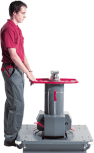 A man operates an industrial tool with a handwheel on a checkered floor, possibly in a workshop setting.
