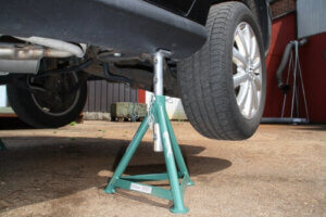 A car is elevated on a green metal jack stand, likely for maintenance, with a building and hose in the background.
