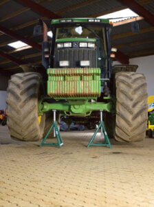 A green tractor is elevated on two metal stands, with its large rear wheels removed, inside a well-lit agricultural maintenance shed.