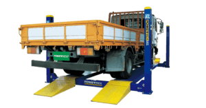 A yellow flatbed truck is elevated on a blue vehicle lift in an apparent maintenance or inspection setup, isolated on a white background.