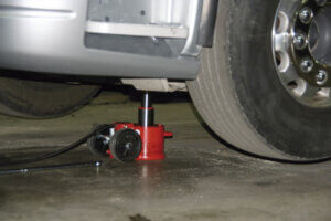 A red hydraulic floor jack is lifting a car's side; tire slightly above ground, on a concrete surface in a garage or workshop setting.