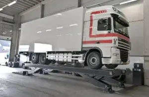 A white and red semi-truck is parked on a vehicle lift inside an industrial garage, suggesting maintenance or inspection.