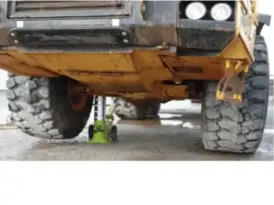 A hydraulic bottle jack supporting the front end of a large, yellow, muddy off-road vehicle with oversized tires, set against a concrete ground.