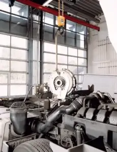 A car's clutch mechanism is being hoisted above the engine bay using a chain pulley in a well-lit, industrial garage setting.