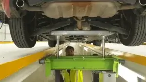 A person in a high-visibility vest peers out from beneath a vehicle on a lift, surrounded by workshop equipment and another car in the background.