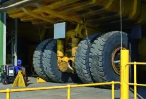 A large yellow mining truck's undercarriage, showing enormous black tires, parked in an industrial maintenance bay with yellow safety railings.