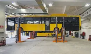 A yellow city bus lifted by orange maintenance stands in an indoor garage with tools and equipment around. Text on bus: "GO FEAST YOUR EYES AT THE ARTS FESTIVAL...GO CREATE...GO ASTRAL AT CARTER OBSERVATORY...GO WELLINGTON".