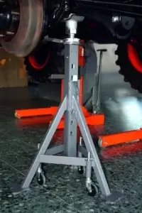 A metal jack stand supports a vehicle's undercarriage, positioned on a glossy floor, with red equipment in the background.