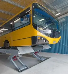 A yellow bus is elevated on a lift inside a maintenance garage, likely undergoing repairs or inspection.