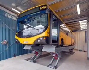 A yellow city bus is elevated on a lift inside a maintenance garage for inspection or repairs.